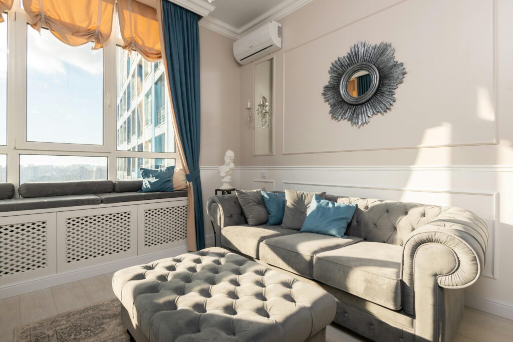 pexels photo 6947272 6947272 Soft couch with cushions near pouf placed at wall with mirror near big window in modern living room with bright sunlight